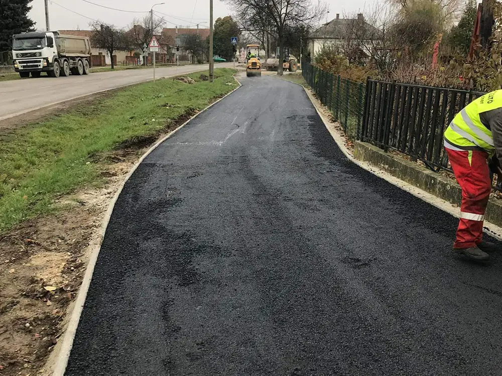 Frissen aszfaltozott utca faluban, munkás láthatósági mellényben, billencs a háttérben