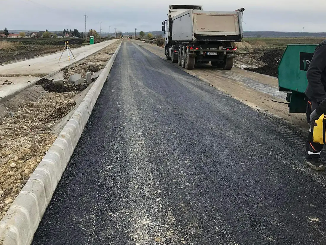 Új építésű út betonszegéllyel, a háttérben billencs; tömörített alap és friss aszfaltréteg