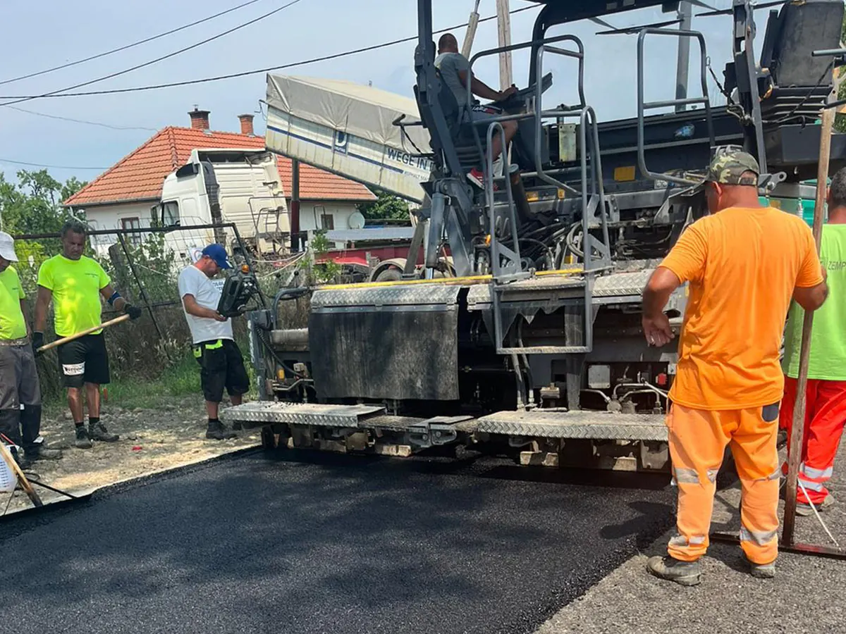 Billencs adagol aszfaltot a terítőgépbe lakóutcában, munkások a gép mellett